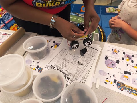 Close-up of a student hand assembling a toy car over worksheets with robot components on a table