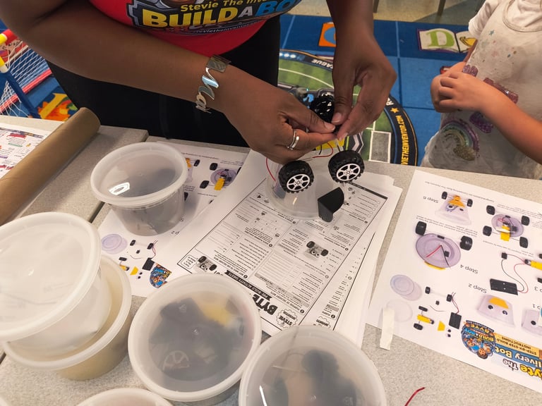 Hands assembling a small toy car above worksheets with purple and yellow components, plastic containers, and instruction sheets on a table