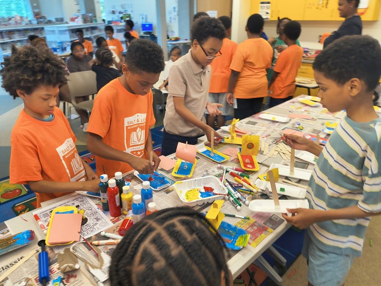 Children in orange shirts and an adult working on a craft project at a community activity table with art supplies.