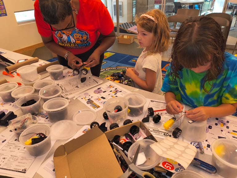 Three children engaged in a colorful art and craft activity with paint supplies and materials spread across a table.
