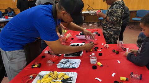 Man in blue shirt building with red LEGO bricks at a table with other people in an indoor workshop setting